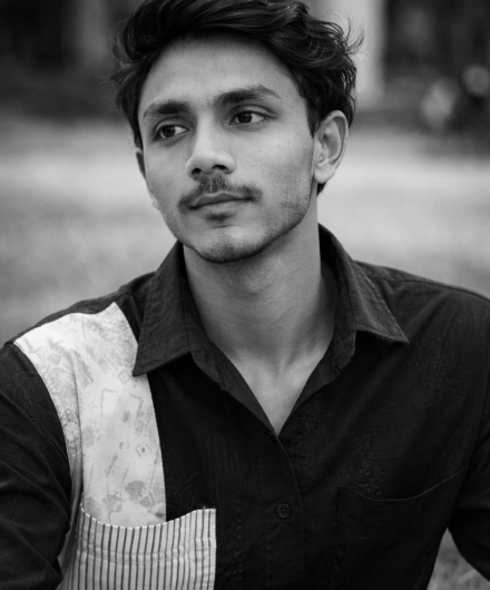 Black-and-white close-up portrait of a young man in a black shirt looking thoughtfully into the distance outdoors.