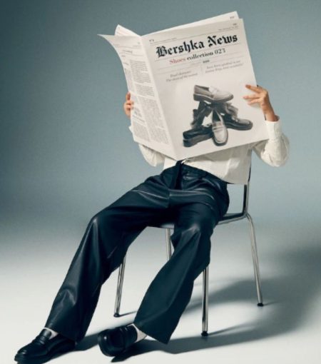 Person sitting on a chair reading a large fashion newspaper while wearing black leather pants and formal shoes in a studio setting.